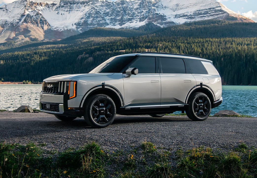 A still shot of the 2027 Kia Telluride Hybrid in silver parked on a rocky lakeside with towering snow-capped mountains and a dense forest in the background, highlighting the driver’s side with bold headlights and all-terrain tires.