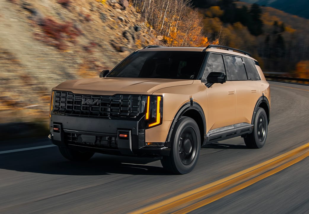 Image of the 2027 Kia Telluride in brown driving on a winding mountain road, showcasing the bold front grille, LED headlights, and rugged SUV design against a backdrop of hills and autumn foliage.
